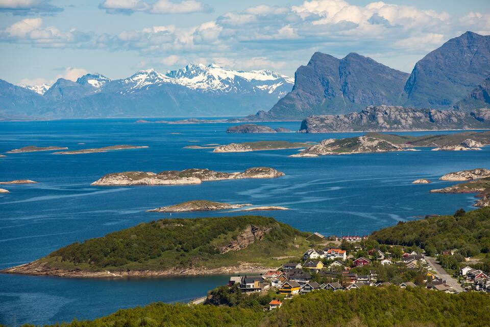 Un pueblo costero con casas, un fiordo y montañas nevadas al fondo.