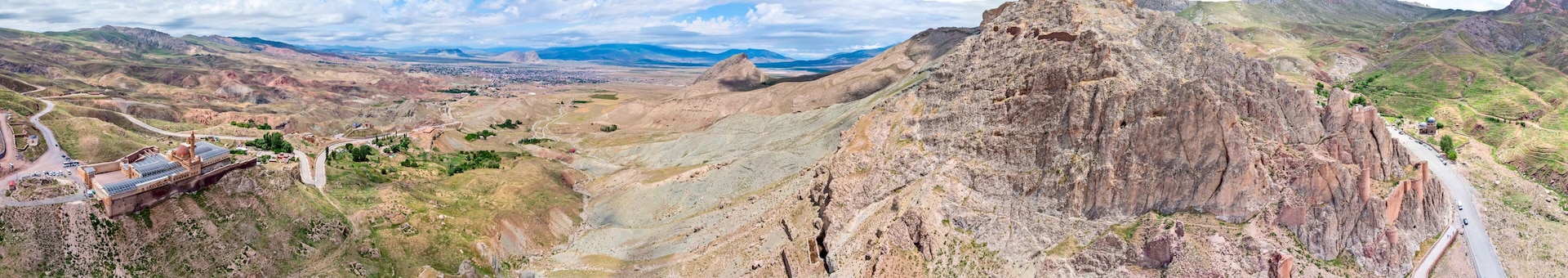 Paisagem montanhosa com um pico rochoso imponente, uma estrada sinuosa e uma cidade ao longe.