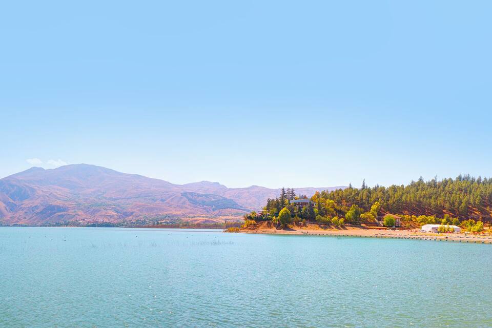 A calm lake with clear turquoise water, surrounded by mountains and a forested area with buildings.