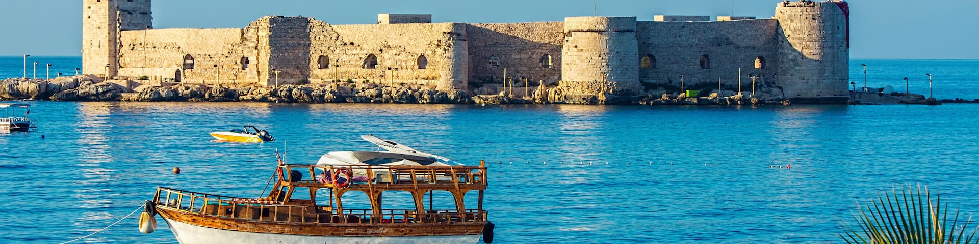 A historic stone fortress by a calm blue sea with a wooden boat anchored nearby.