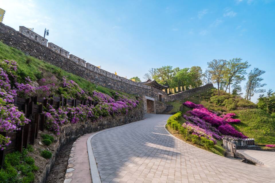 Un chemin de pierres serpentant à travers un jardin où poussent des fleurs violettes, le tout bordé d’un mur de pierres.