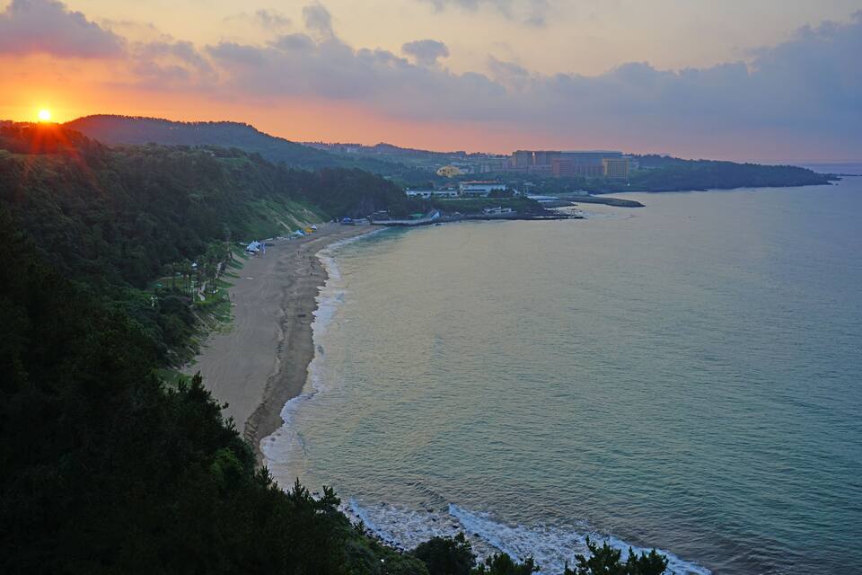 A coastal landscape with a beach, ocean, and hills during sunset.