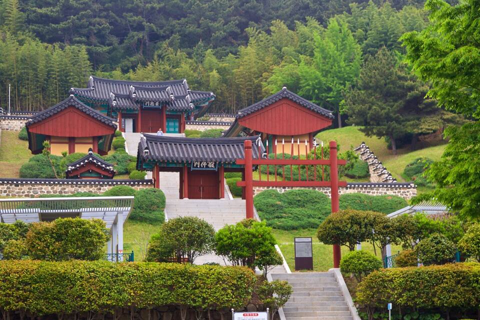 Traditional Korean architecture with red buildings and grey tiled roofs, surrounded by lush greenery and trees.