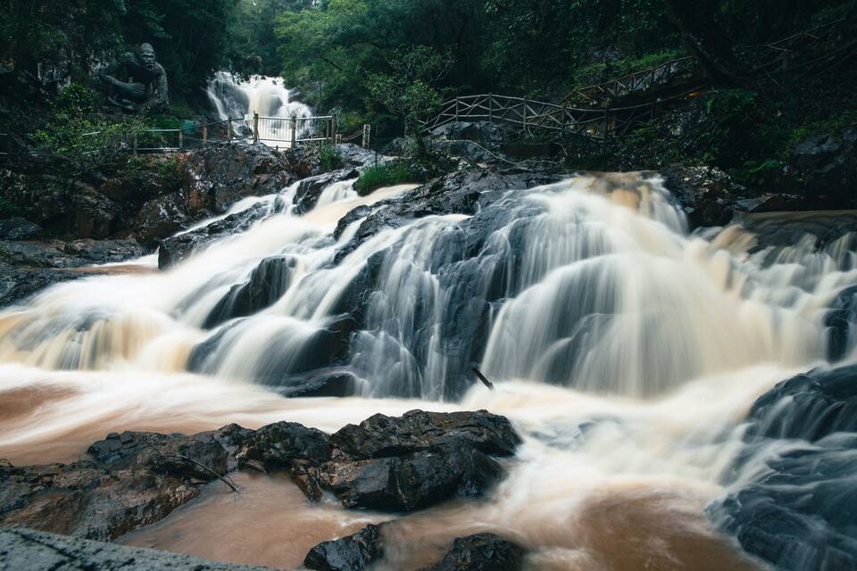 Ein Wasserfall stürzt über Felsen, im Hintergrund eine Holzbrücke und eine Statue.