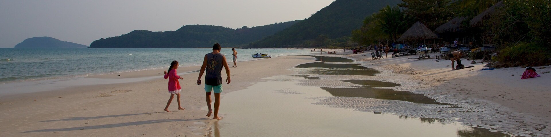 Uma praia de areia com pessoas caminhando, mar calmo e colinas ao fundo.