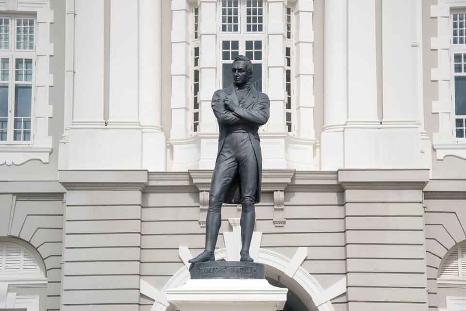 Una estatua de Stamford Raffles frente a un edificio blanco con ventanas de arco y columnas.