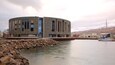 Un bâtiment moderne à la conception circulaire, situé près d’une étendue d’eau calme, entouré d’une côte rocheuse et dominé par un paysage montagneux.