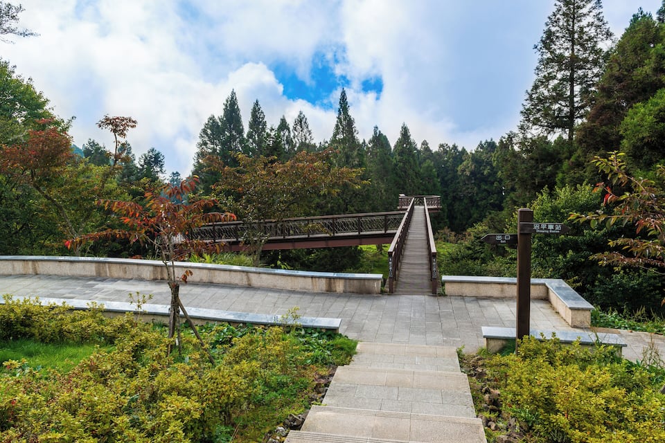 A stone pathway leading to a wooden bridge over a body of water, surrounded by lush greenery and trees.