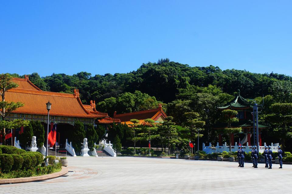 Um edifício tradicional chinês com telhado vermelho, rodeado por vegetação exuberante e estátuas.