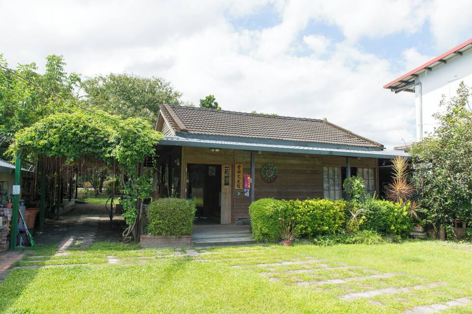 A traditional wooden building with a tiled roof, surrounded by lush greenery and a stone pathway leading to the entrance.