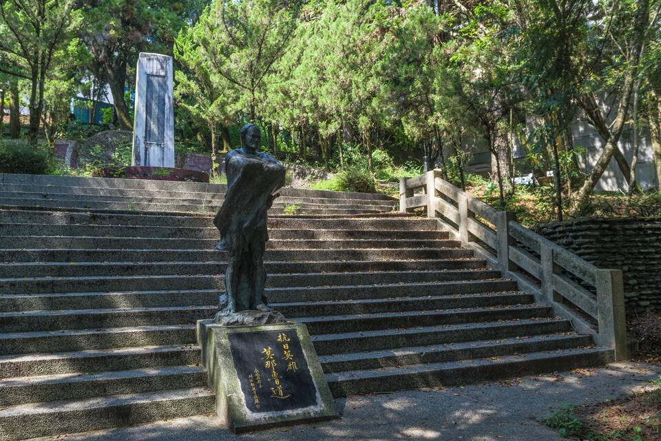 A statue on a stone staircase with a plaque, surrounded by trees.