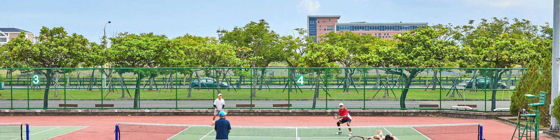 Una cancha de tenis con jugadores, árboles y un edificio al fondo.