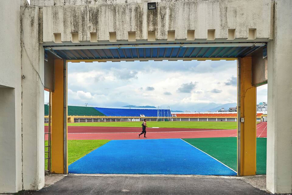 Un estadio de atletismo con pista azul, carriles verdes y rojos, y una persona corriendo.