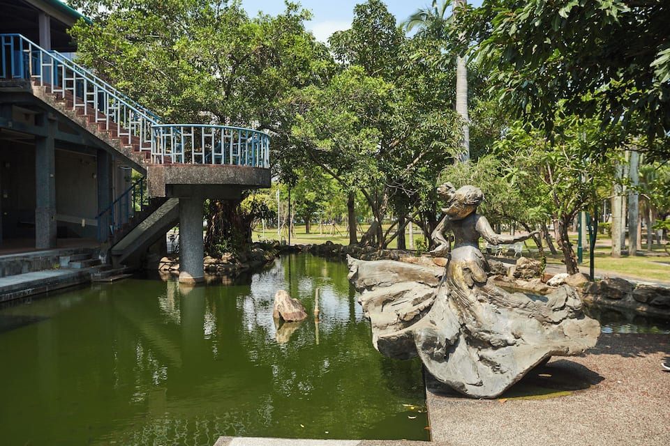 Um parque com lago, uma estátua e uma escadaria que leva a um edifício.