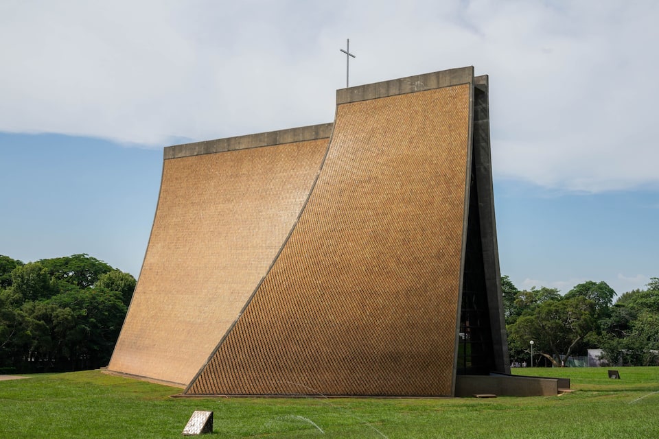 A large, modern building with a sloped roof and a cross on top, surrounded by a grassy area and trees.