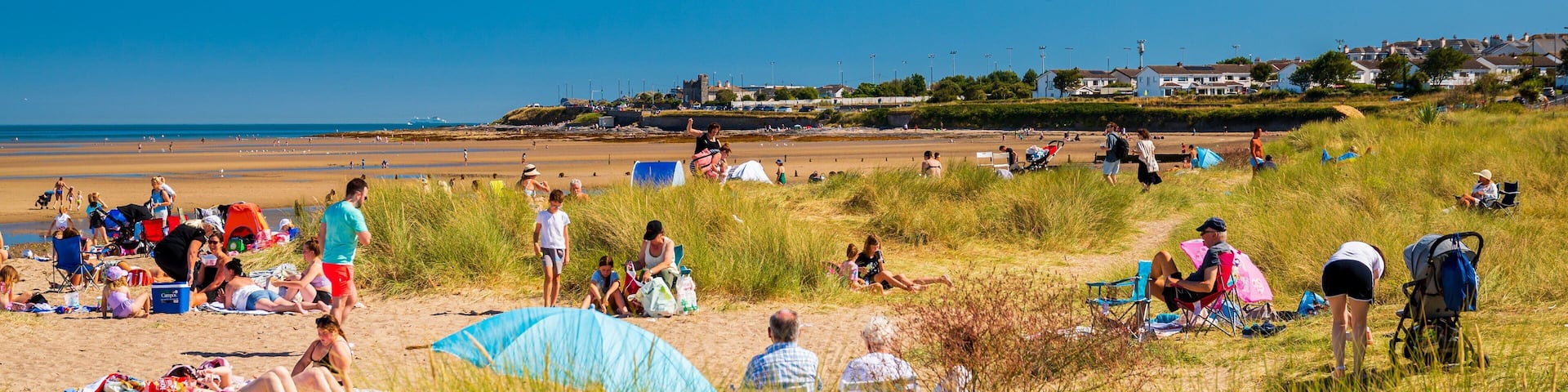 Une plage où l’on voit des gens se prélasser au soleil et se promener, des dunes herbeuses et, au loin, des maisons.