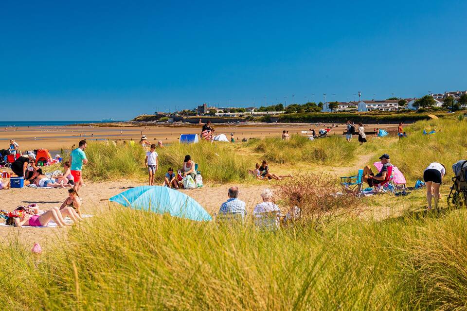 En strand med folk, der solbader og går, græsbevoksede klitter og huse i baggrunden.