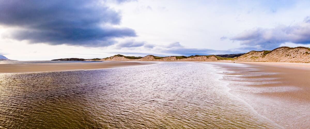 A sandy beach with gentle waves, dunes, and a cloudy sky.