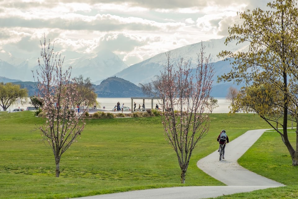 A park with a paved path, trees, a lake, and mountains in the background.