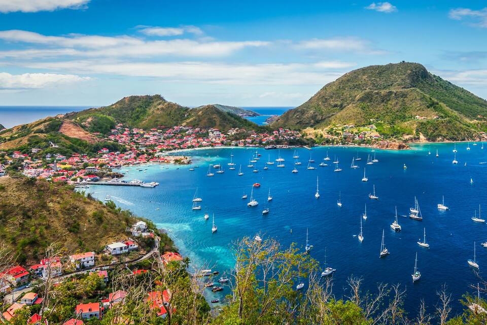 A coastal town with red-roofed buildings, a bay filled with sailboats, and a prominent hill in the background.