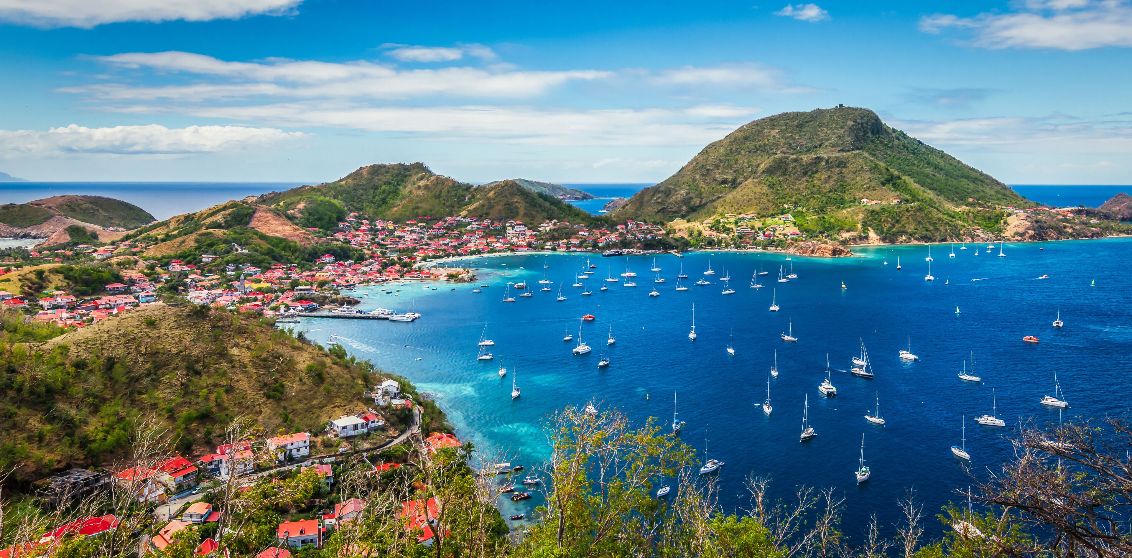 A coastal town with red-roofed buildings, a bay filled with sailboats, and a prominent hill in the background.