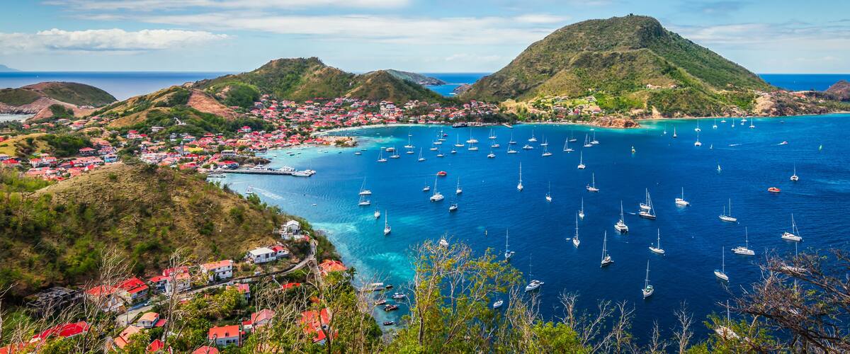 A coastal town with red-roofed buildings, a bay filled with sailboats, and a prominent hill in the background.
