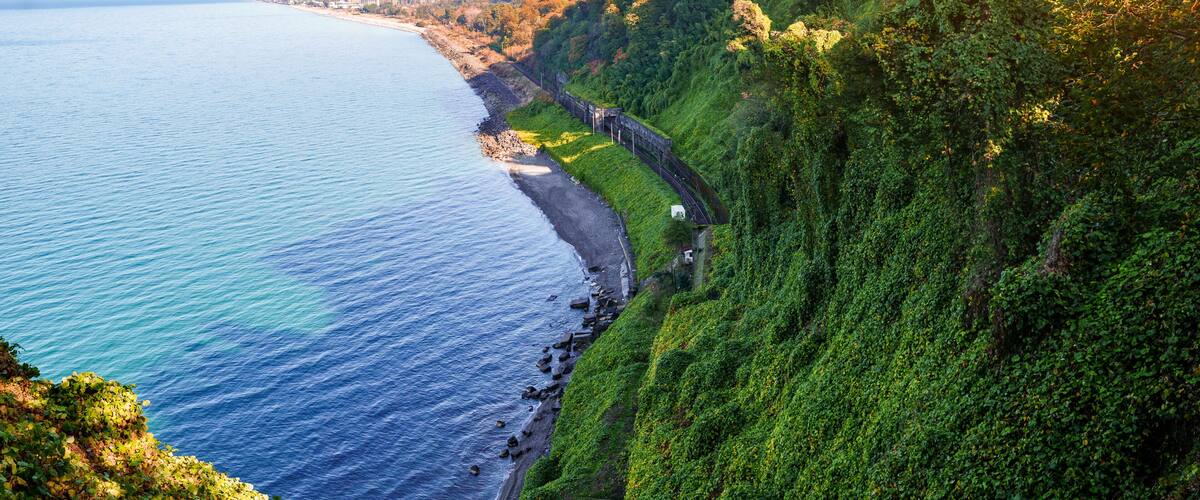 A coastal landscape with a beach, dense greenery, and a clear view of the ocean.