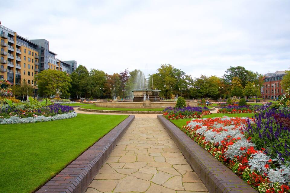 Ein angelegter Garten mit einem zentralen Brunnen, umgeben von Blumenbeeten und einem gepflasterten Weg.