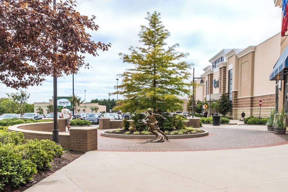 A circular plaza with a bronze statue, a large tree, and a JOANN store in the background.
