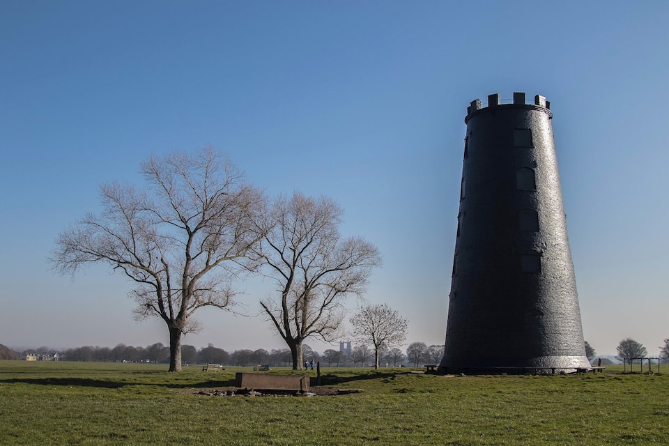 Un'alta torre cilindrica in un parco con alberi spogli e cielo azzurro intenso.