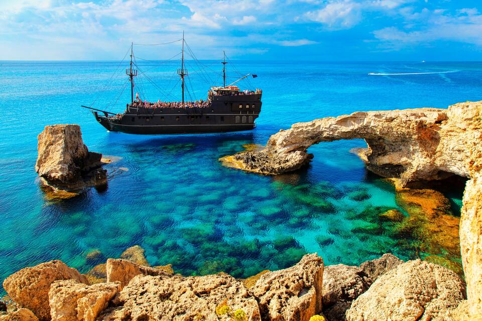 A ship anchored near a natural rock arch in clear blue waters.