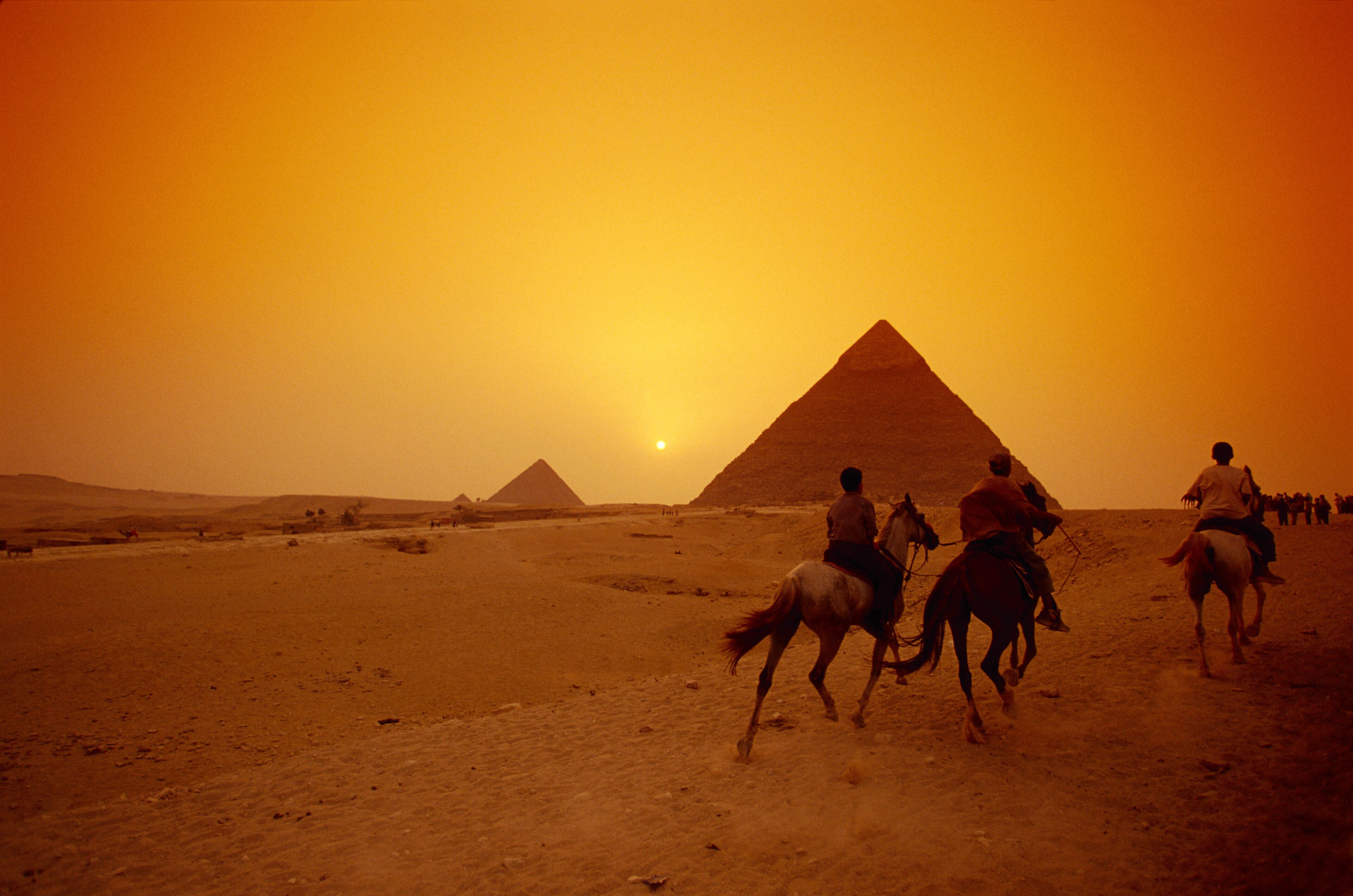 A desert landscape with pyramids and people riding horses.