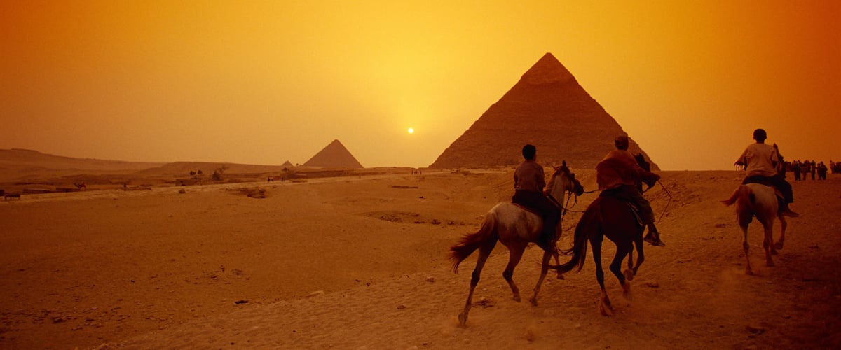 A desert landscape with pyramids and people riding horses.