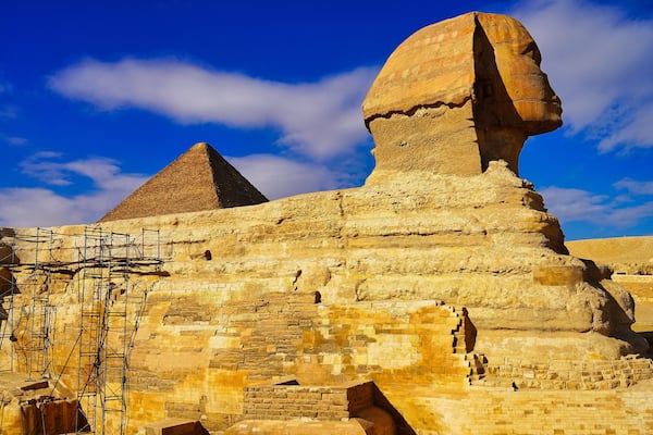 A grand view of the Great Sphinx of Giza with scaffolding for restoration work on a bright hot day on the Giza plateau with the Pyramid of Khufu in the background