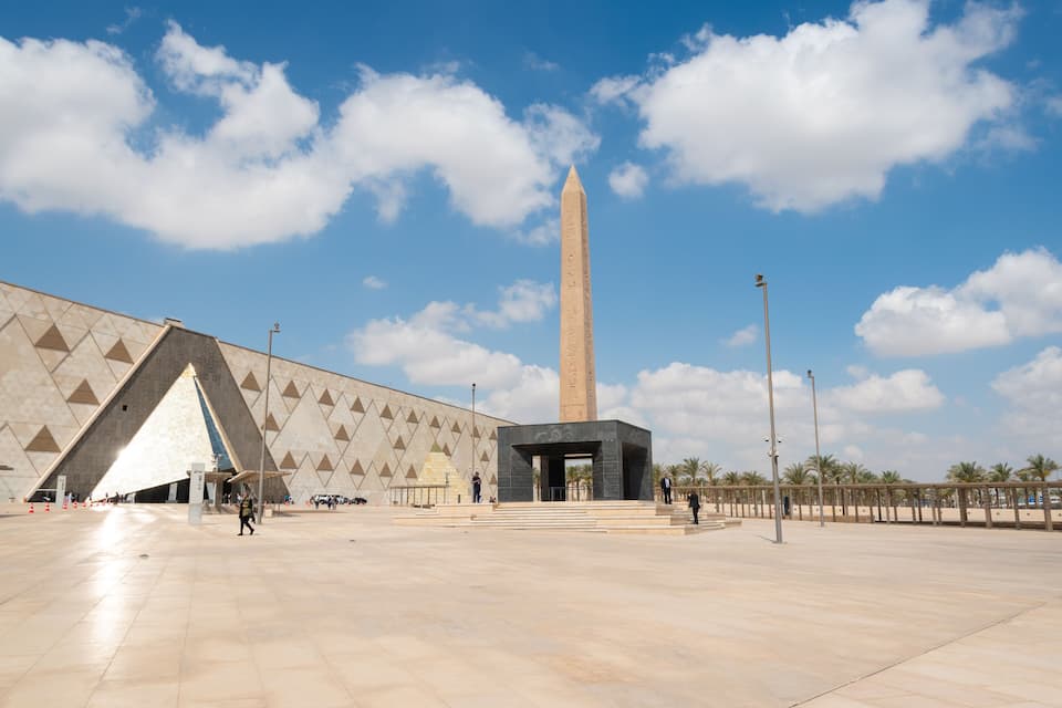 Een grote obelisk, een modern gebouw met een geometrisch gevelontwerp en een helderblauwe lucht met wolken.