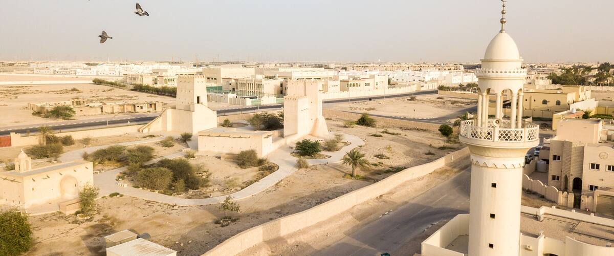 A mosque with a tall minaret, surrounded by a desert landscape with sparse vegetation and a few buildings in the background.