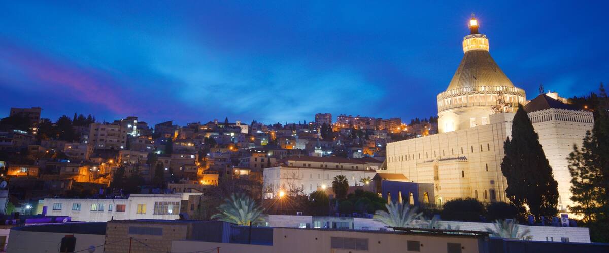 A cityscape at dusk with a prominent mosque featuring a large dome and minarets, surrounded by residential buildings and palm trees.