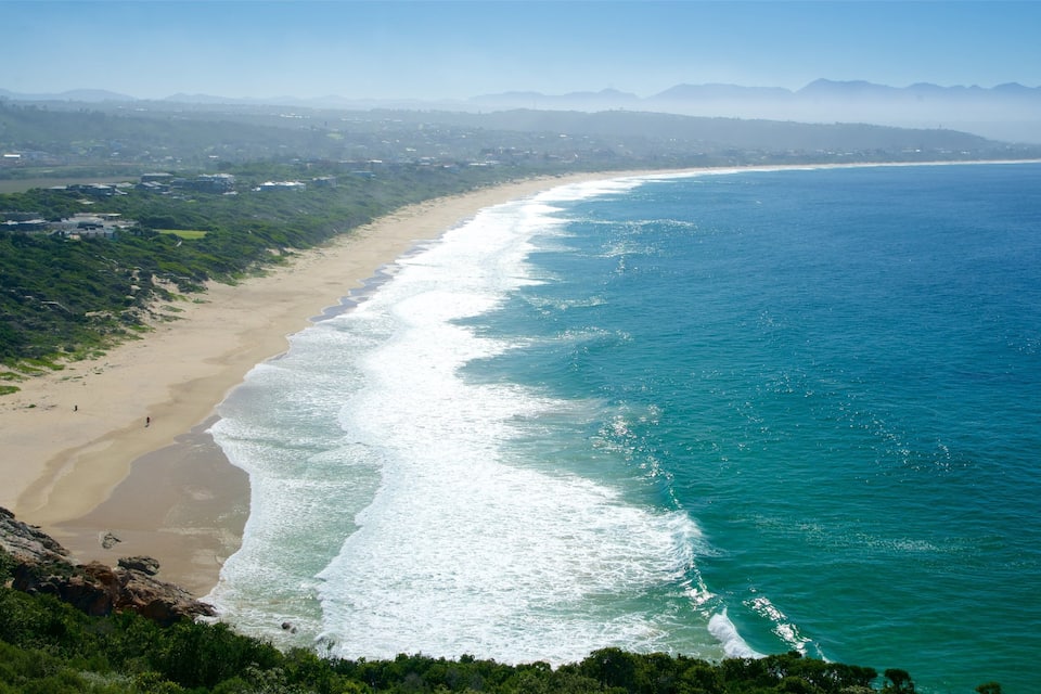 Paisagem costeira com praia arenosa, ondas oceânicas azul-turquesa e montanhas verdes ao fundo.