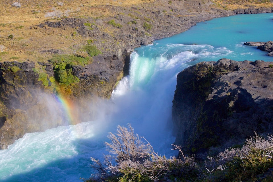 A waterfall with a rainbow, surrounded by rocky terrain and sparse vegetation.