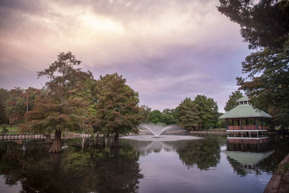 Un parc tranquil amb un estany que reflecteix, un pavelló i una font.
