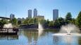 Ein Park mit einem See, zwei Springbrunnen, einem hölzernen Steg und einer Stadtansicht im Hintergrund.