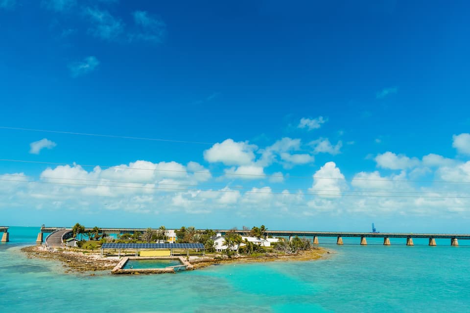 A coastal bridge, turquoise waters, and a small island with buildings and solar panels.