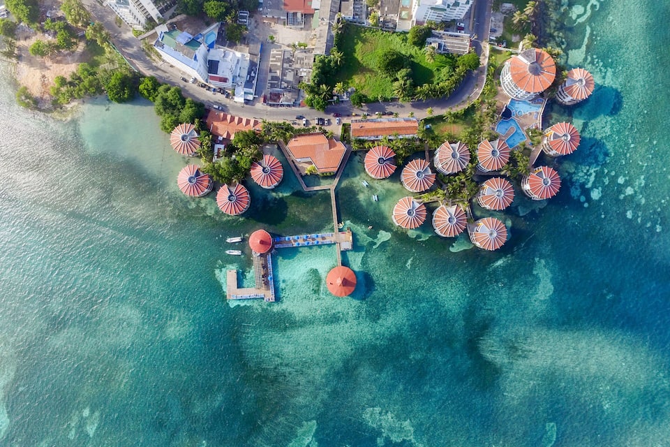 Vista aérea de un complejo turístico costero con varias zonas a la sombra de sombrillas, un muelle y edificios circundantes.