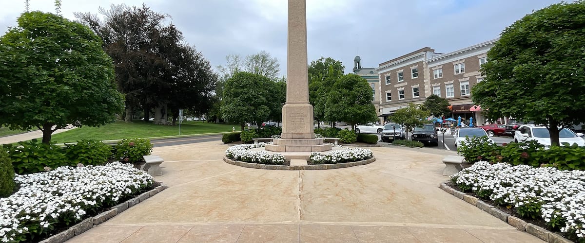 A tall monument in a landscaped square with surrounding trees and a building in the background.