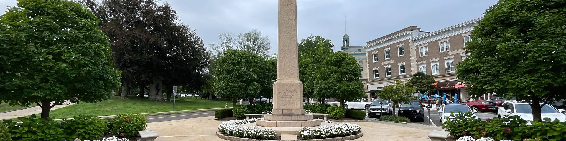 A tall monument in a landscaped square with surrounding trees and a building in the background.