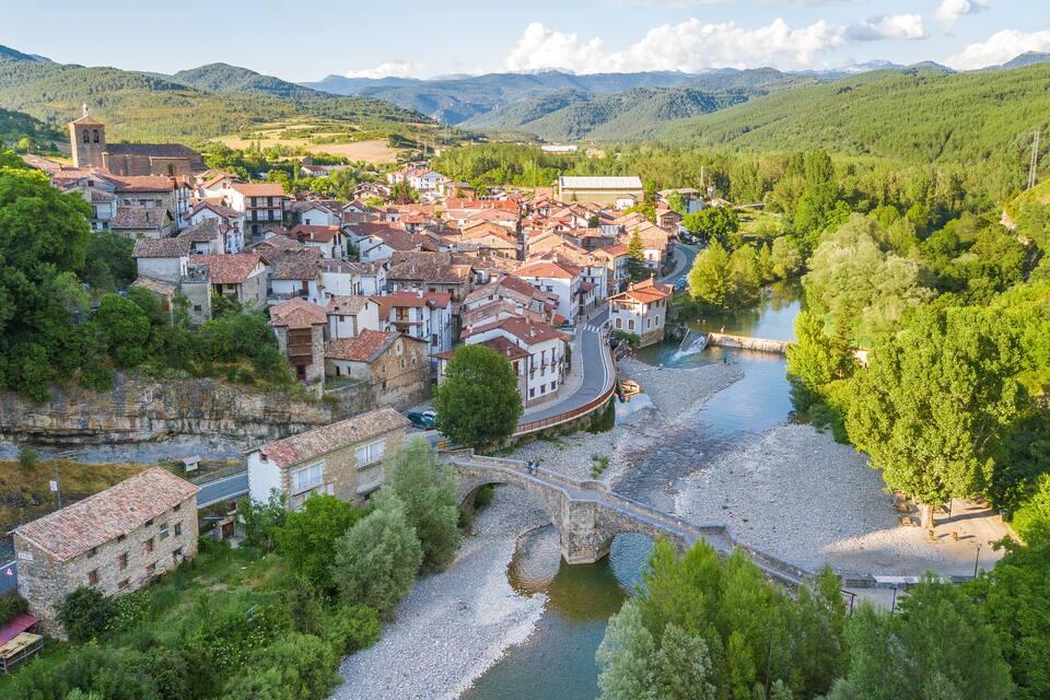 A town with red-tiled roofs, a river, and surrounding hills.