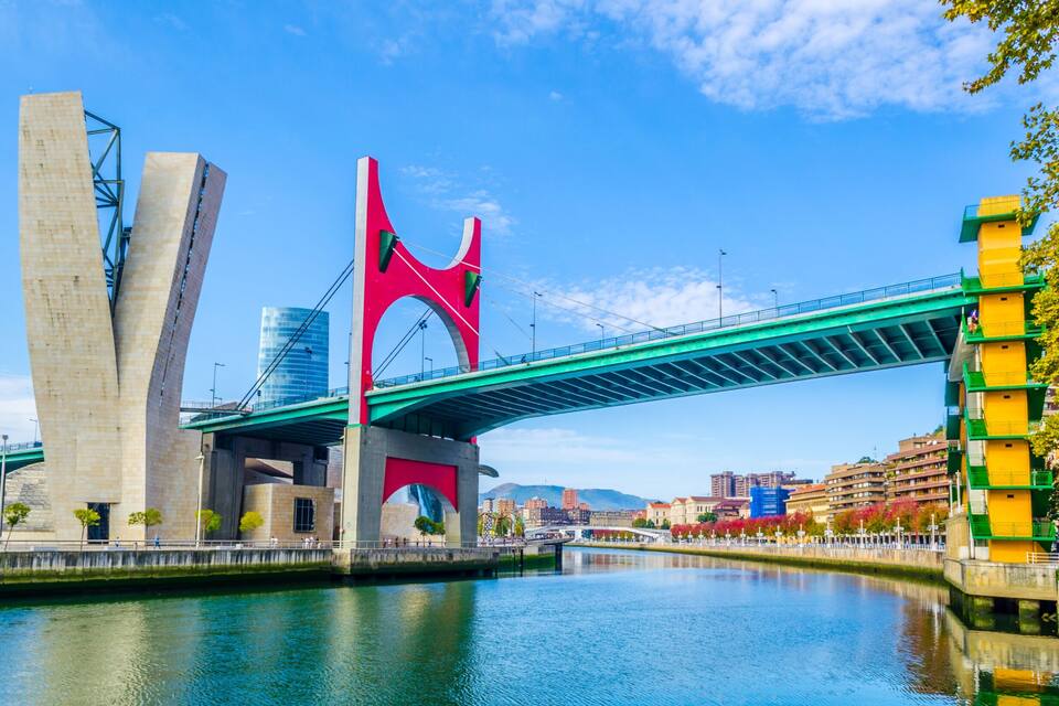 A modern bridge with a distinctive red arch and green deck spanning a river, with buildings and trees in the background.