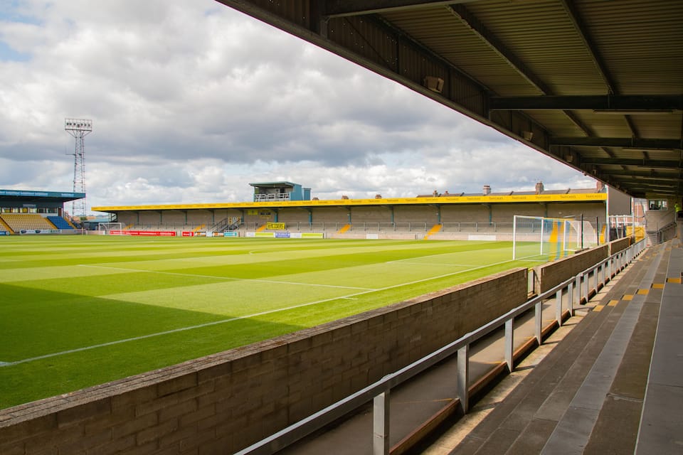 A football stadium with a green pitch, yellow and blue seating, and a covered spectator area.