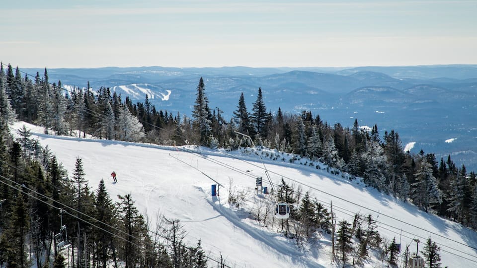 A snowy mountain slope with a ski lift, trees, and distant mountains.