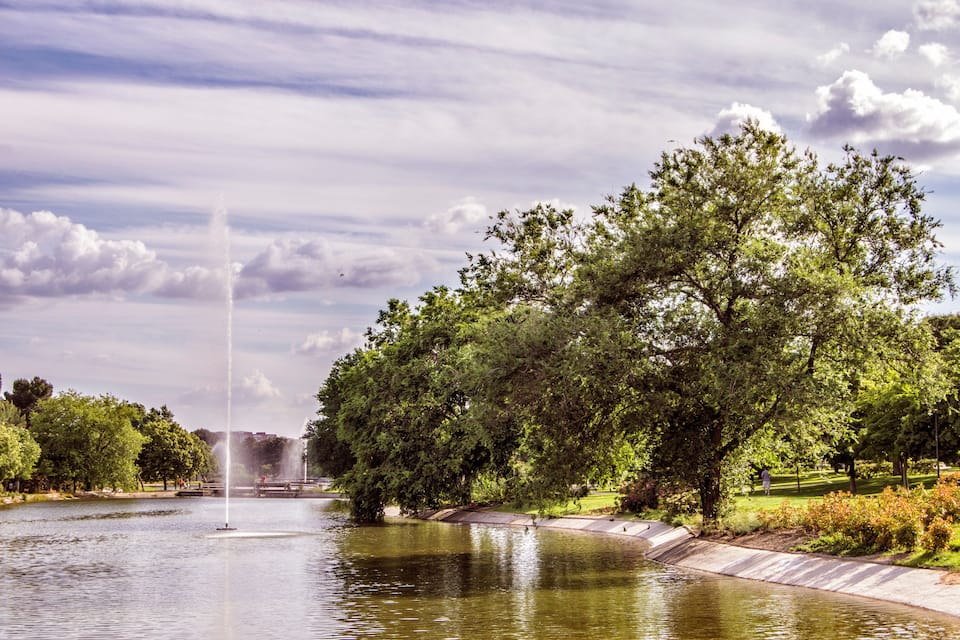 A park with a fountain, trees, and a calm body of water.